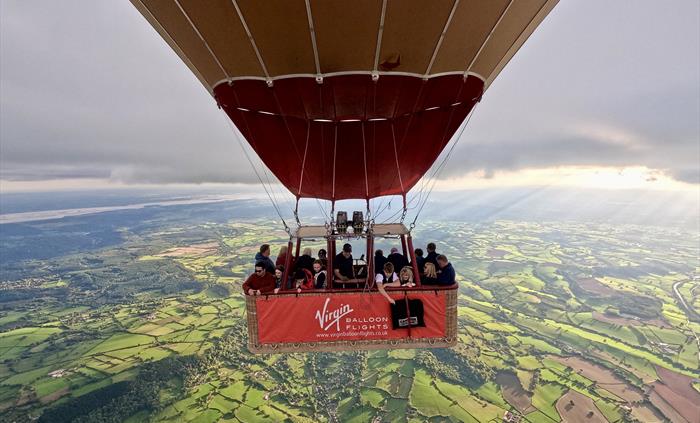 Passengers enjoying a Virgin Balloon Flights hot air balloon ride over the Welsh countryside, with patchwork fields and sunlit views below.